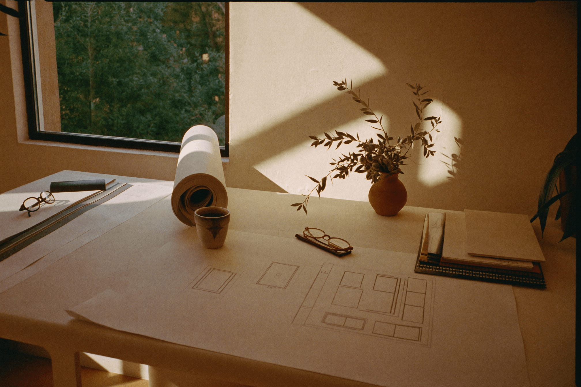 A sunlit studio desk with plans, samples and an olive sprig.